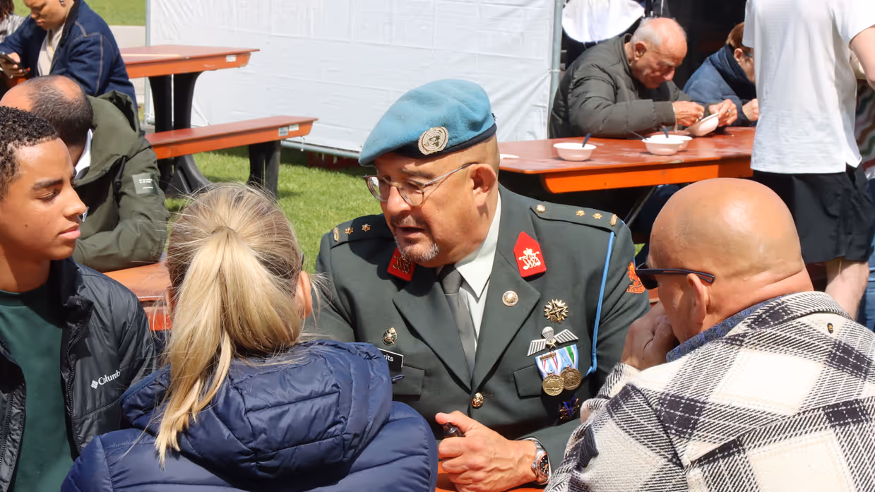 Een militair in uniform met een blauwe baret zit aan een tafel. Hij is in gesprek met jongeren. 
