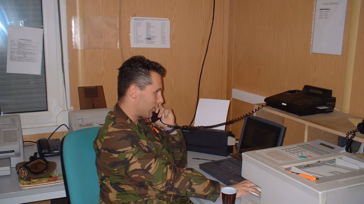 Een oude foto van Marcel Pelster waarbij hij achter een bureau met een fax aan de telefoon zit.