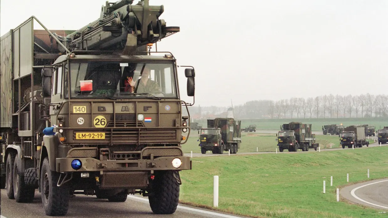 Een colonne militaire vrachtwagens met Patriot-materieel rijdt over een weg. Voorop rijdt een DAF-vrachtwagen met een antennemast, gevolgd door meerdere voertuigen met ondersteunende apparatuur en lanceerinrichtingen.