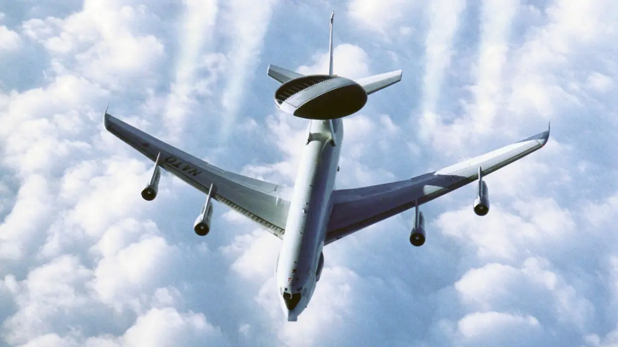 Bovenaanzicht van een AWACS boven een bewolkte lucht. De motoren laten vier condensstrepen achter. 