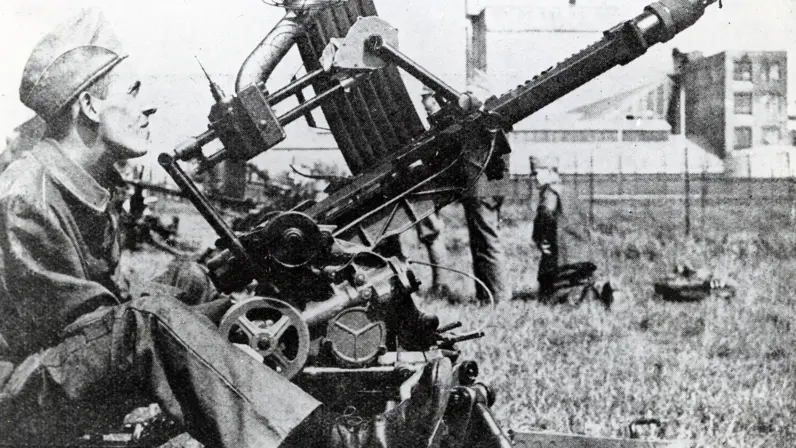 Een soldaat in uniform bedient een zware, oude machinegeweeropstelling in een open veld met gras en gebouwen op de achtergrond. 