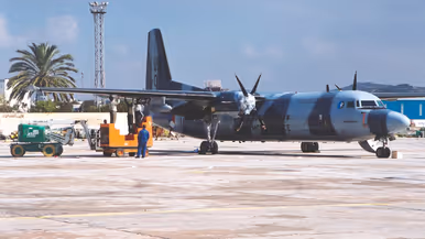  Links: Een groot, blauw-wit militair vliegtuig op een vliegveld. Mensen en voertuigen laden spullen in. Palmbomen en een toren op de achtergrond, op een zonnige, heldere dag. Rechts: Twee personen plakken zwarte letters op de onderkant van een vliegtuigvleugel. Het vliegtuig heeft de tekst 'UN' en cijfers op de zijkant.