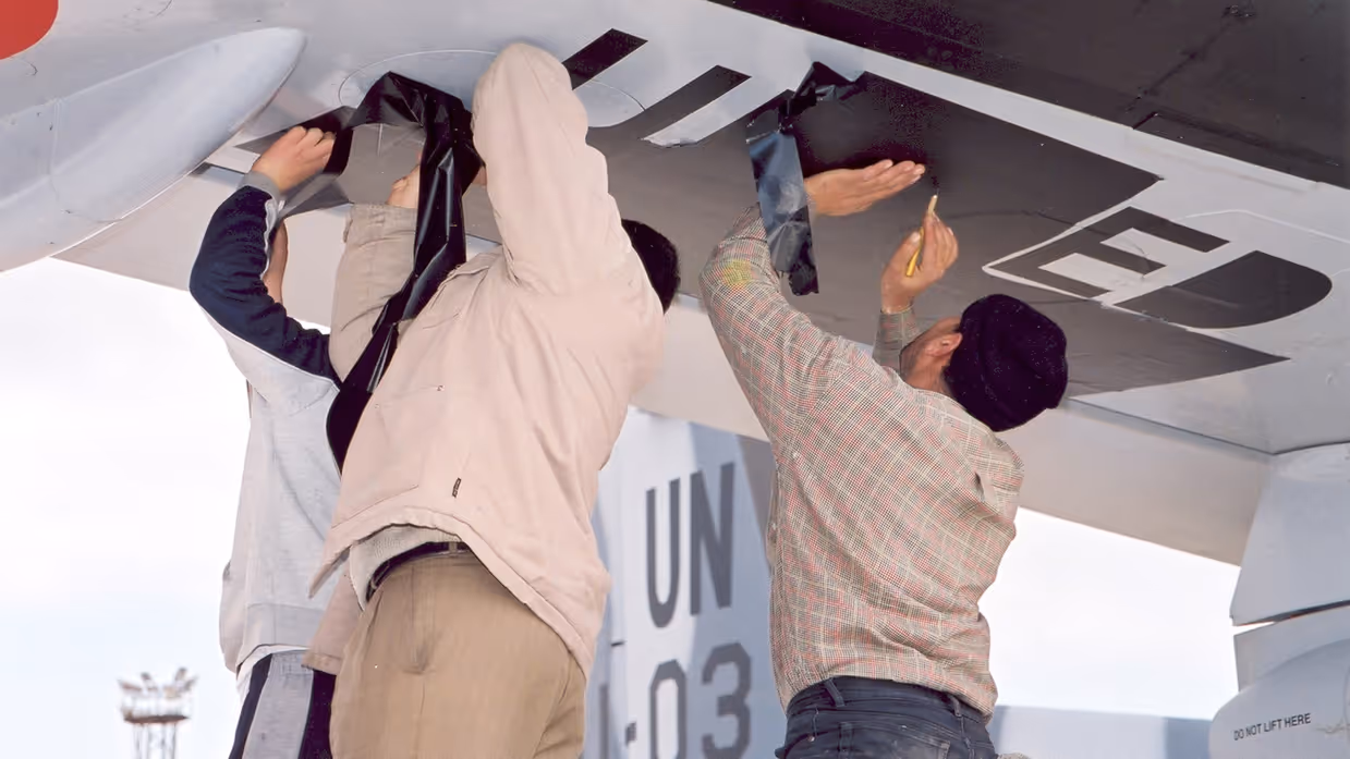  Links: Een groot, blauw-wit militair vliegtuig op een vliegveld.  Mensen en voertuigen laden spullen in. Palmbomen en een toren op de achtergrond, op een zonnige, heldere dag. Rechts: Twee personen plakken zwarte letters op de onderkant van een vliegtuigvleugel.Het vliegtuig heeft UN en cijfers op de zijkant.