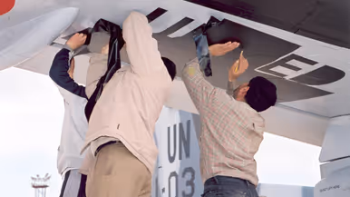  Links: Een groot, blauw-wit militair vliegtuig op een vliegveld.  Mensen en voertuigen laden spullen in. Palmbomen en een toren op de achtergrond, op een zonnige, heldere dag. Rechts: Twee personen plakken zwarte letters op de onderkant van een vliegtuigvleugel.Het vliegtuig heeft UN en cijfers op de zijkant.