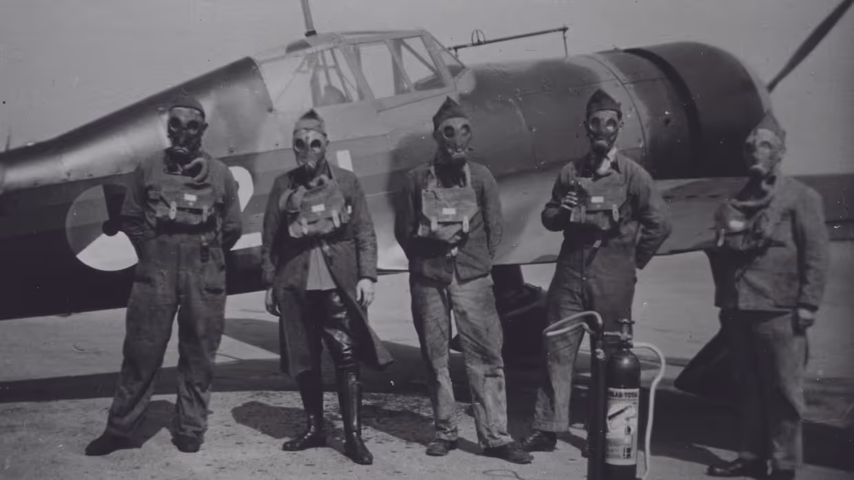 Vijf mannen in vliegtenue met gasmaskers en veldmasker staan voor een oud militair vliegtuig, met een brandblusapparaat naast hen. De foto is zwart-wit.
