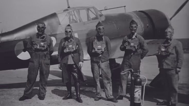 Vijf mannen in vliegtenue met gasmaskers en veldmasker staan voor een oud militair vliegtuig, met een brandblusapparaat naast hen. De foto is zwart-wit.