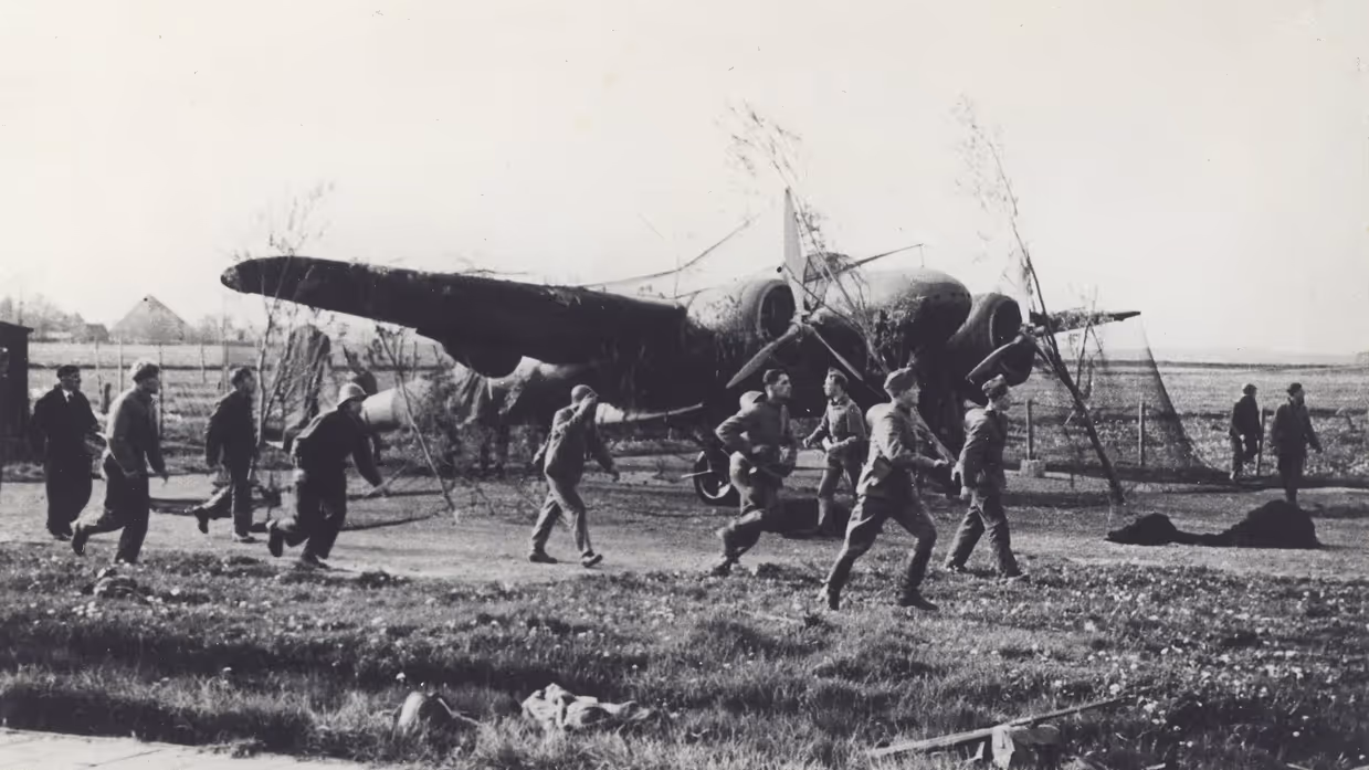 Mannen lopen over een grasveld naar een neergestort vliegtuig met zichtbare schade en een open landschap op de achtergrond.