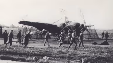 Mannen lopen over een grasveld naar een neergestort vliegtuig met zichtbare schade en een open landschap op de achtergrond.