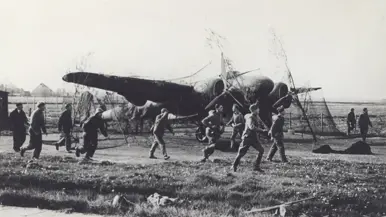 Mannen lopen over een grasveld naar een neergestort vliegtuig met zichtbare schade en een open landschap op de achtergrond.