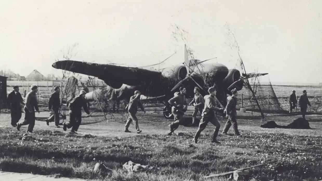 Mannen lopen over een grasveld naar een neergestort vliegtuig met zichtbare schade en een open landschap op de achtergrond.