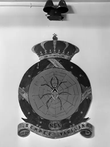Zwart-witfoto van een embleem met een spin in een web en een lauwerkrans eromheen.
