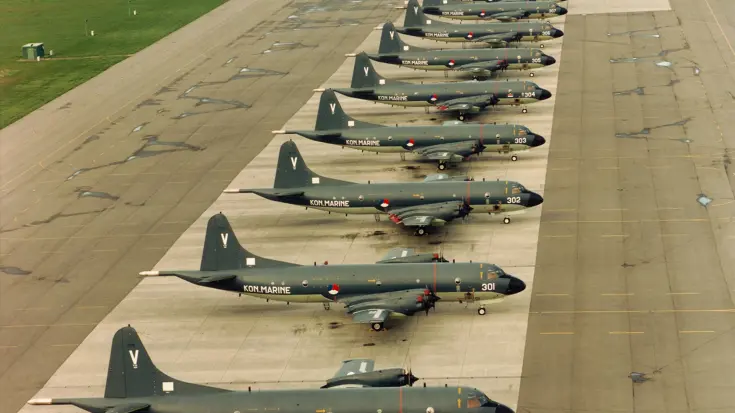 Een line-up van alle 13 Lockheed P-3C (Update II) Orion lange afstand maritieme patrouillevliegtuigen (1981-2005) op het platform van Marinevliegkamp Valkenburg.  Alttekst: Dertien donkere maritieme patrouillevliegtuigen van het type Orion staan netjes op een rij op een startbaan.