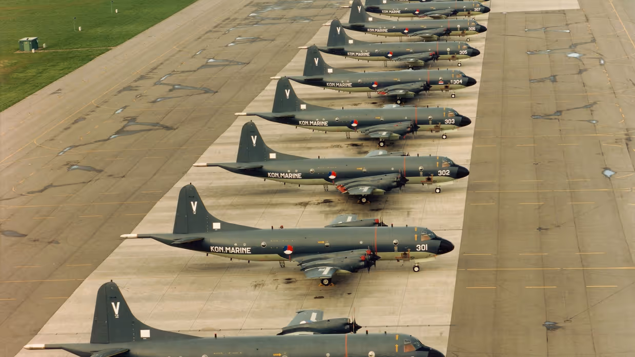 Een line-up van alle 13 Lockheed P-3C (Update II) Orion lange afstand maritieme patrouillevliegtuigen (1981-2005) op het platform van Marinevliegkamp Valkenburg.  Alttekst: Dertien donkere maritieme patrouillevliegtuigen van het type Orion staan netjes op een rij op een startbaan.