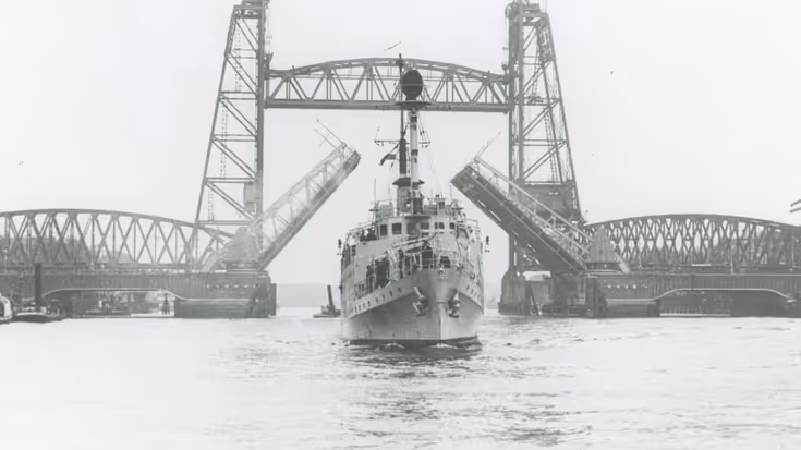 Een groot schip vaart onder een hoge, metalen ophaalbrug door. De brug is open en heeft twee torens met een roosterachtige constructie boven het water.