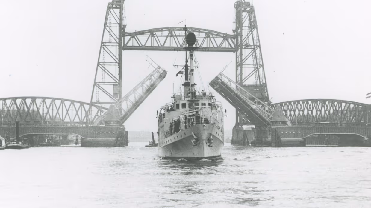 Een groot schip vaart onder een hoge, metalen ophaalbrug door. De brug is open en heeft twee torens met een roosterachtige constructie boven het water.