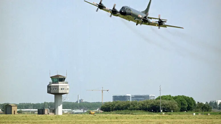 Een vliegtuig vliegt laag over een groen veld met een verkeerstoren en gebouwen op de achtergrond onder een bewolkte lucht.