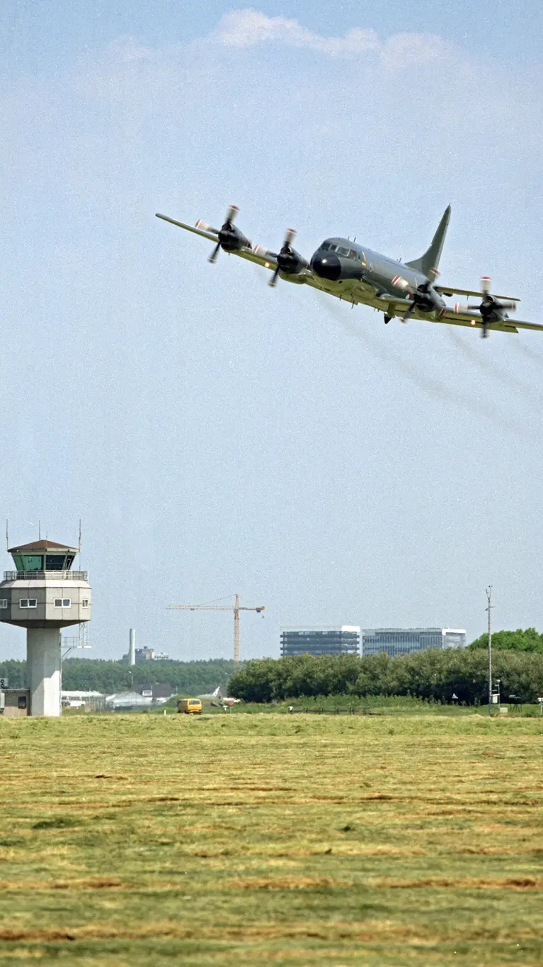 Een vliegtuig vliegt laag over een groen veld met een verkeerstoren en gebouwen op de achtergrond onder een bewolkte lucht.