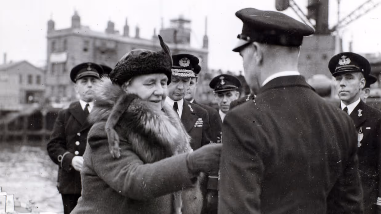 Links: Een vrouw in een dikke jas met bontkraag is omringd door mannen in uniformen in een haven. Rechts: Een metalen medaille met een krans, een "W" in het midden, en een kroontje erboven, aan een verticaal gestreept lint.