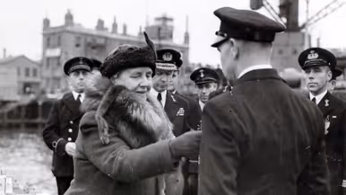 Links: Een vrouw in een dikke jas met bontkraag is omringd door mannen in uniformen in een haven. Rechts: Een metalen medaille met een krans, een "W" in het midden, en een kroontje erboven, aan een verticaal gestreept lint.