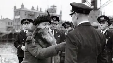 Links: Een vrouw in een dikke jas met bontkraag is omringd door mannen in uniformen in een haven. Rechts: Een metalen medaille met een krans, een "W" in het midden, en een kroontje erboven, aan een verticaal gestreept lint.