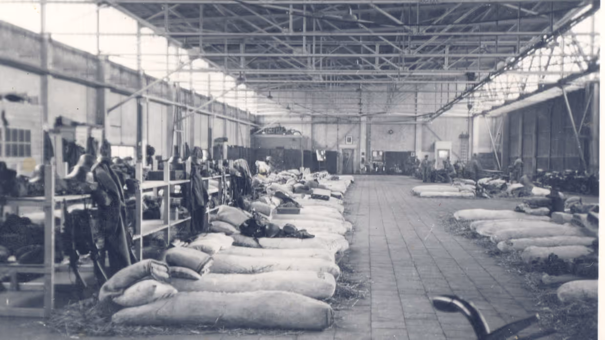 Een grote, open loods met rijen zakken met stro op de grond, metaalframes, en gereedschappen aan de zijkanten, waar de opgeroepen reservisten moeten slapen.