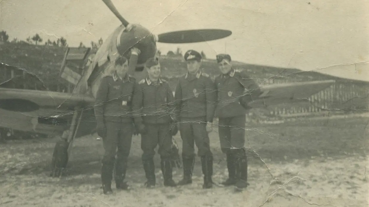 4 mannen staan in uniform voor een vliegtuig met een grote propeller op een open veld met bomen.