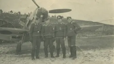 4 mannen staan in uniform voor een vliegtuig met een grote propeller op een open veld met bomen.