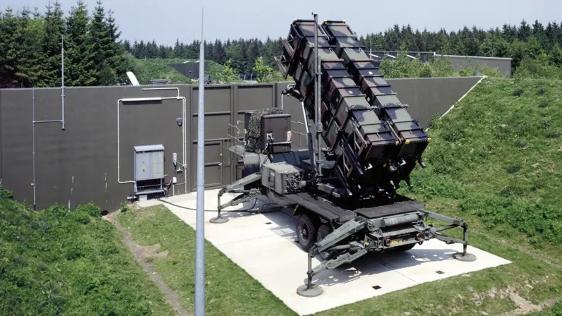 Een Patriot-lanceerinrichting staat opgesteld op een militair terrein in Duitsland. Het lanceersysteem staat op een verharde opstelplaats, omgeven door een betonnen beveiliging en groene wallen.