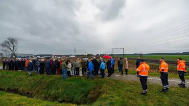 Links een groep mensen, waaronder militairen in uniform bij een bord langs de spoorlijn. Rechts: bloemen en kransen bij een bord met daarop een afbeelding van een oude trein, terwijl een moderne trein langs raast.