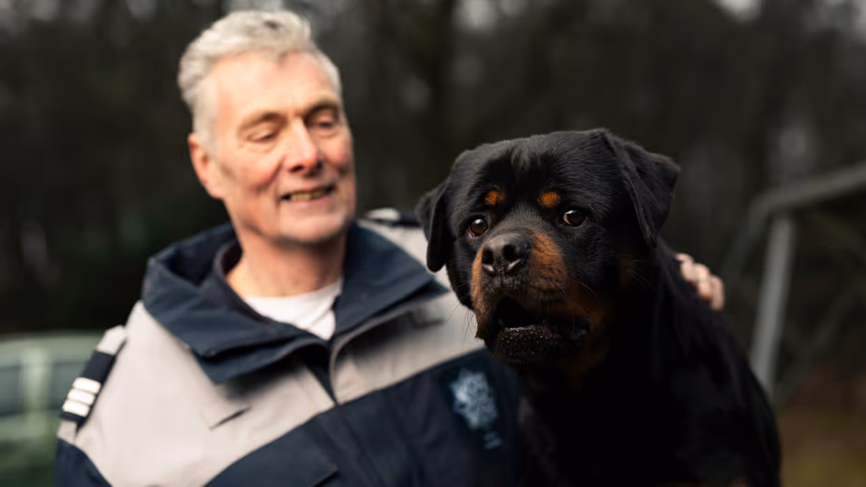 Links een man in uniform. Hij omarmt een grote zwart-bruine hond met opgetrokken oren. Achtergrond toont een bosrijke omgeving met een metalen hek. Rechts een glimlachende man in uniform staat naast een zwarte Rottweiler met open bek. Bosrijke omgeving op de achtergrond, met een ontspannen en vriendelijke sfeer. 