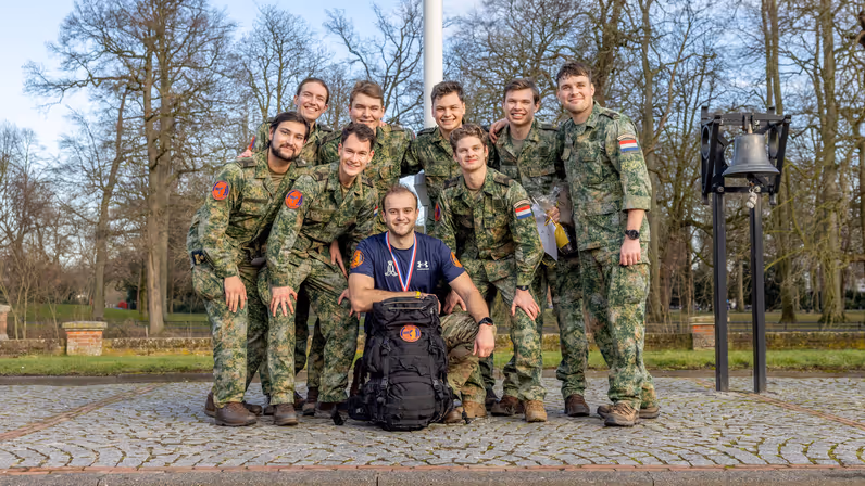 Zeven mannen in militaire kleding staan glimlachend in een groep buiten, met bomen op de achtergrond. Een man knielt voor hen met een grote rugzak en een medaille om. 