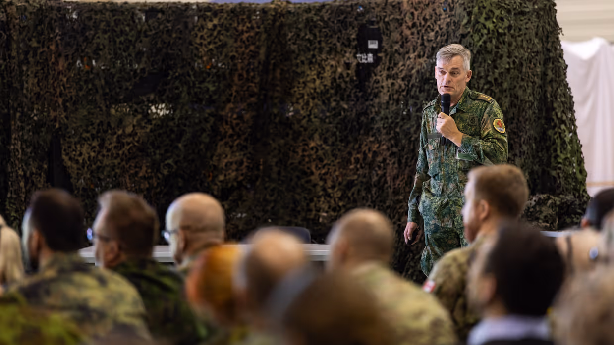  Links: Een militair in uniform staat voor een groen gecamoufleerd doek en spreekt met een microfoon. Een groep mensen in uniform en burgerkleding luistert aandachtig. Rechts: Een bord met het embleem "Casualty Move 2026 Medical Quadrigga 2026" staat op de voorgrond. Achterin zitten mensen in een grote ruimte naar een presentatie te kijken.