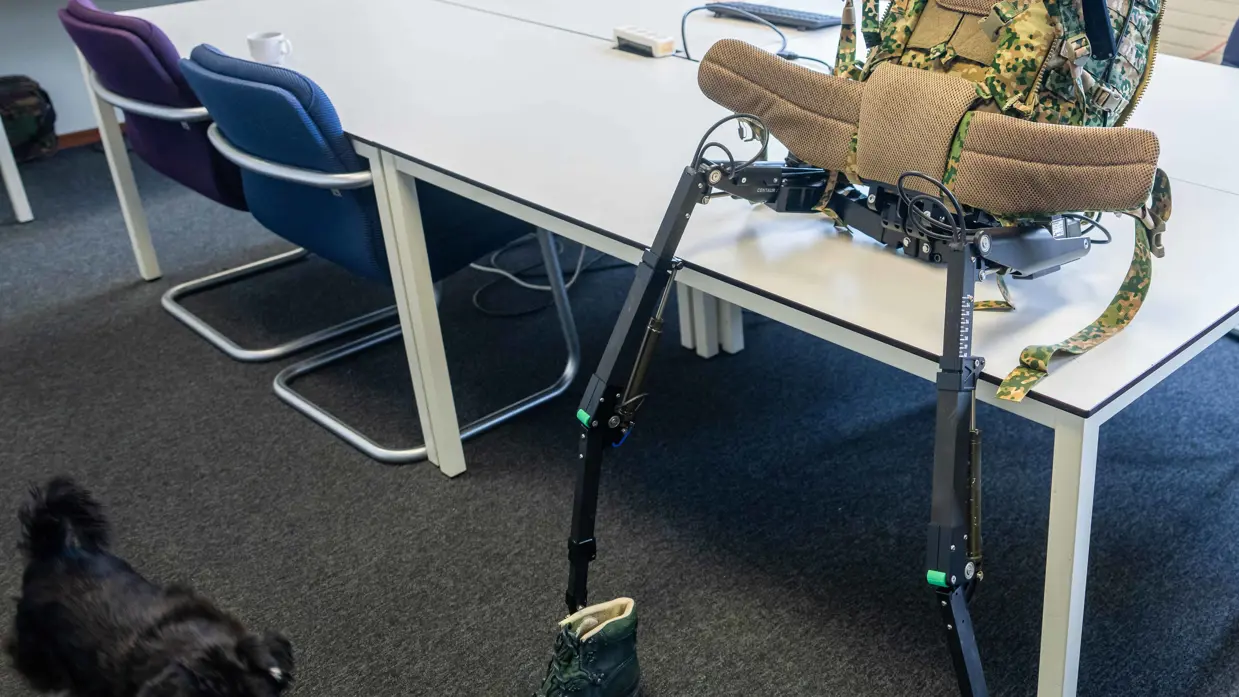 Een exoskelet met camouflage rugzak en groene bergschoenen staat op een tafel in een klaslokaal met stoelen en tafels.