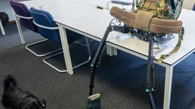 Links: een exoskelet met camouflage rugzak en groene bergschoenen staat op een tafel in een klaslokaal met stoelen en tafels.    Rechts een groene bergschoen met een zwarte metalen meetlat ernaast staat op een grijze vloer, tweede schoen op de achtergrond. 
