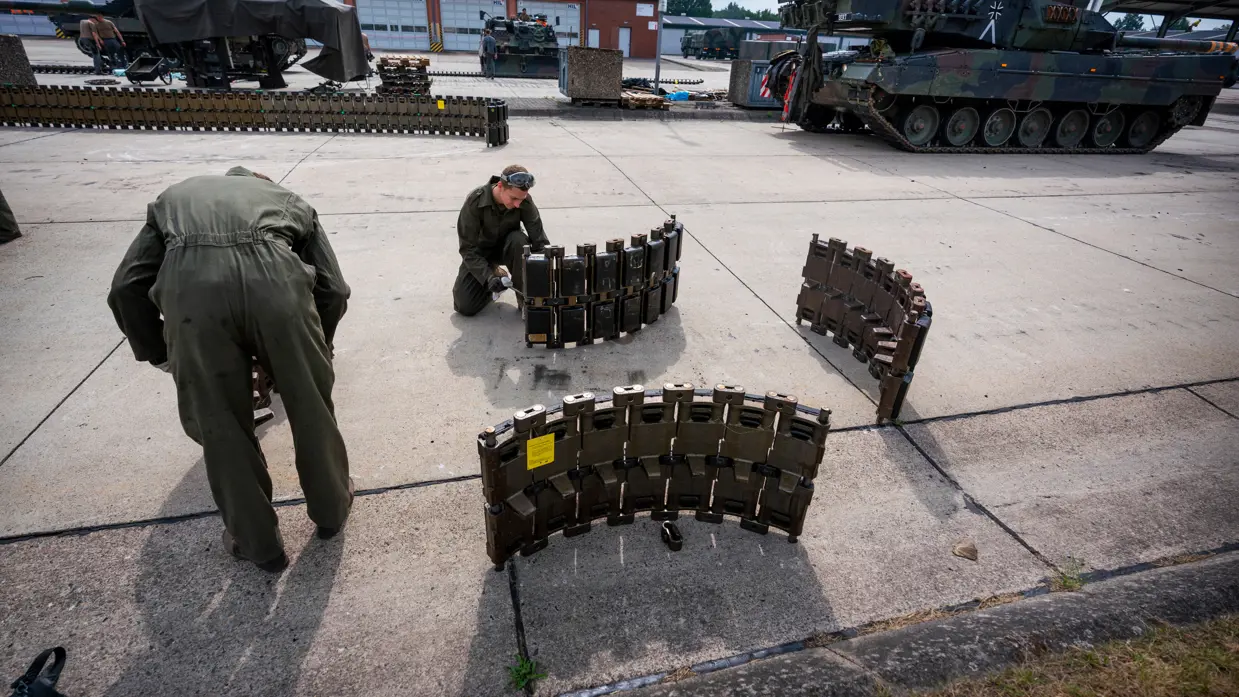 Twee soldaten in groene overall monteren een zware, metalen structuur op een betonnen oppervlak met tanks op de achtergrond.