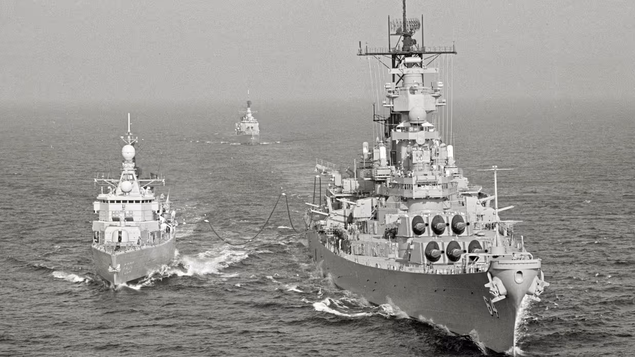  Drie oorlogsschepen varen op ruwe zee: twee grote schepen voorop, verbonden door kabels, en een kleiner schip op de achtergrond.