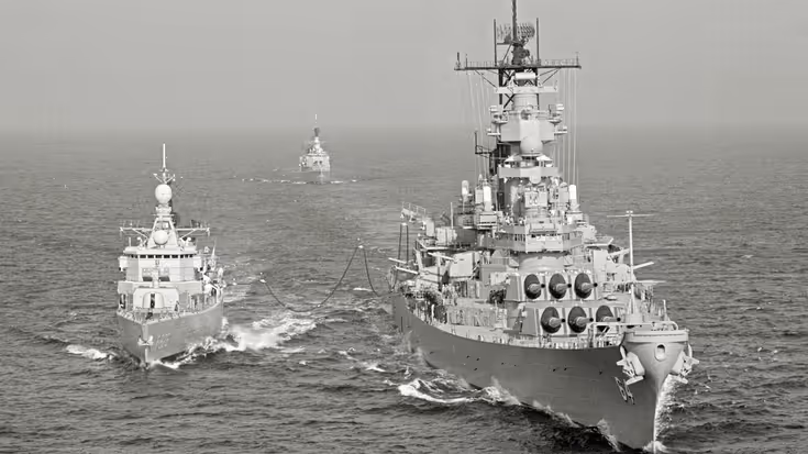  Drie oorlogsschepen varen op ruwe zee: twee grote schepen voorop, verbonden door kabels, en een kleiner schip op de achtergrond.