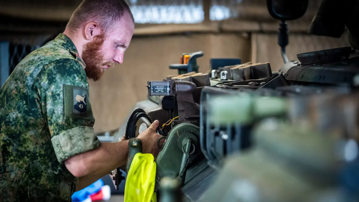 Een militair in camouflage-uniform werkt geconcentreerd aan een mechanisch onderdeel in een werkplaats met gereedschap.
