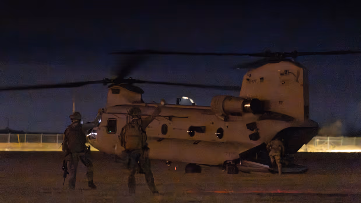  In schemering stapt laatste militair via de achterkant de heli in. Twee militairen op het terrein geven met gebalde vuist de vlieger een teken.
