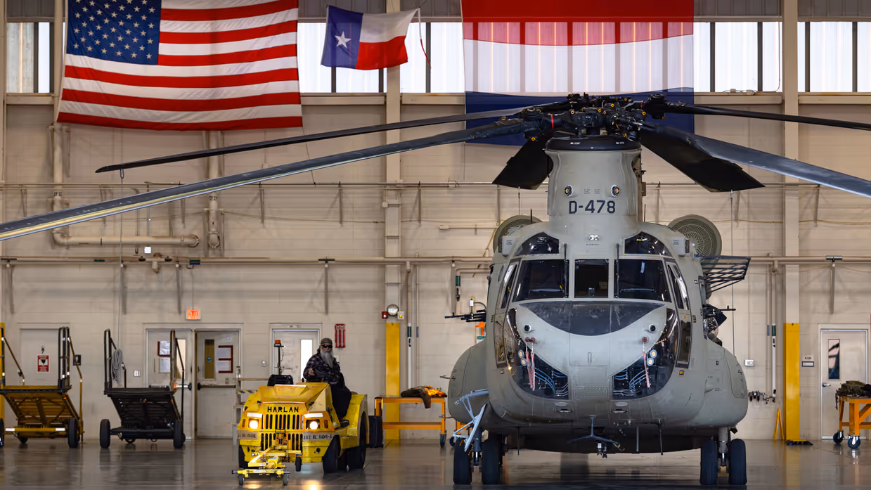 Chinook in een hangaar met erboven aan de muur een Amerikaanse en een Nederlandse vlag. Naast de helikopter een geel onderhoudsvoertuig met daarop een Amerikaanse inhuurkracht.