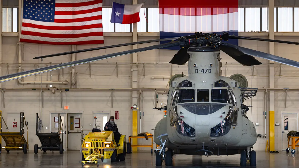 Chinook in een hangaar met erboven aan de muur een Amerikaanse en een Nederlandse vlag. Naast de helikopter een geel onderhoudsvoertuig met daarop een Amerikaanse inhuurkracht.