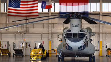 Chinook in een hangaar met erboven aan de muur een Amerikaanse en een Nederlandse vlag. Naast de helikopter een geel onderhoudsvoertuig met daarop een Amerikaanse inhuurkracht.