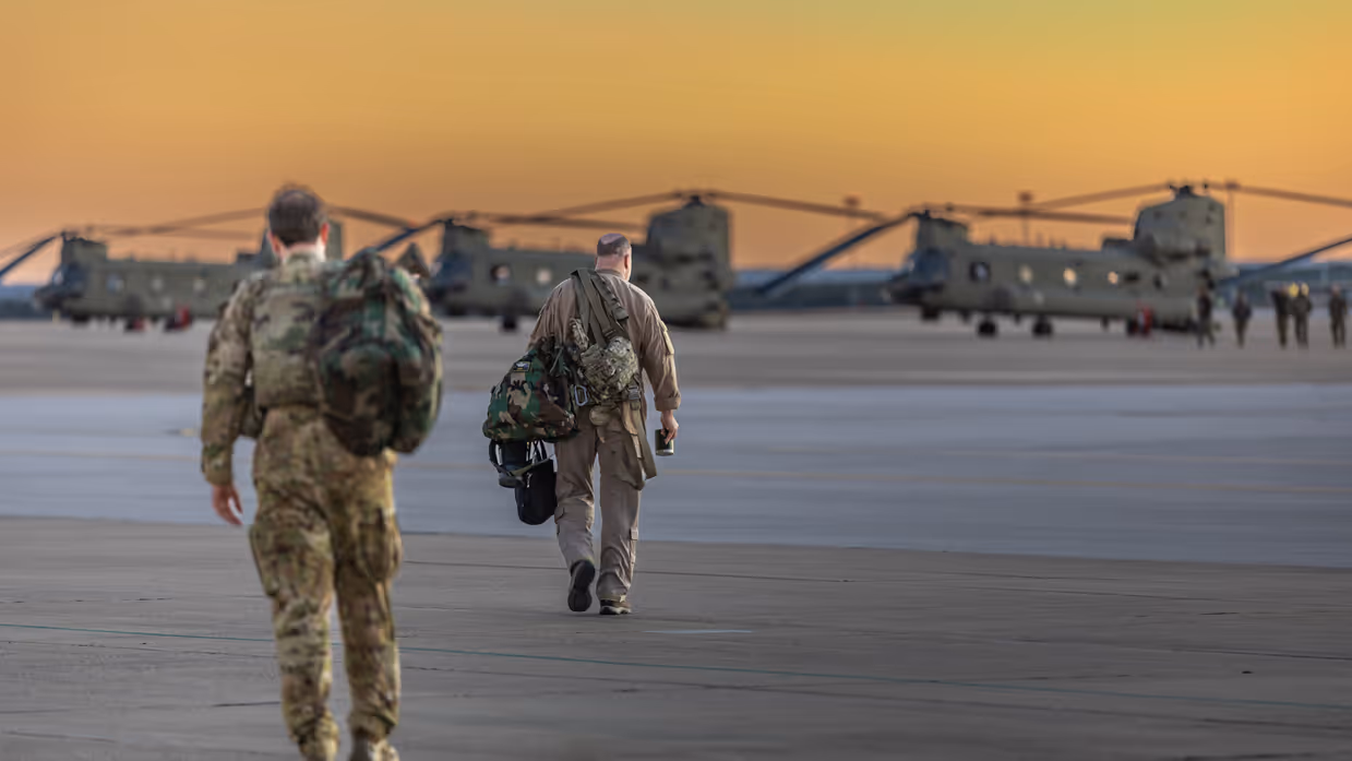Twee militairen lopen met tassen richting drie Chinooks op een platform. Op de achtergrond een oranje-gele lucht.