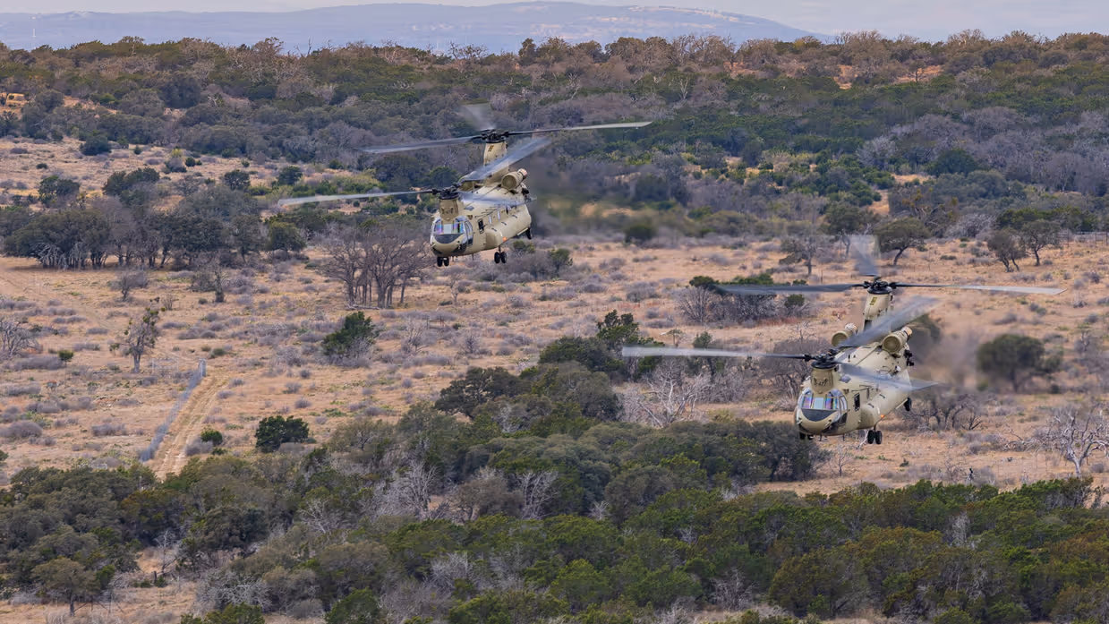 Twee laagvliegende Chinooks boven uitgestrekt landschap met zand en plukken begroeiing. Op de achtergrond heuvels.