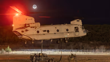 Een militair voertuig wordt bij duisternis onder een Chinook gehangen op een veld. Erboven schijnt de maan.