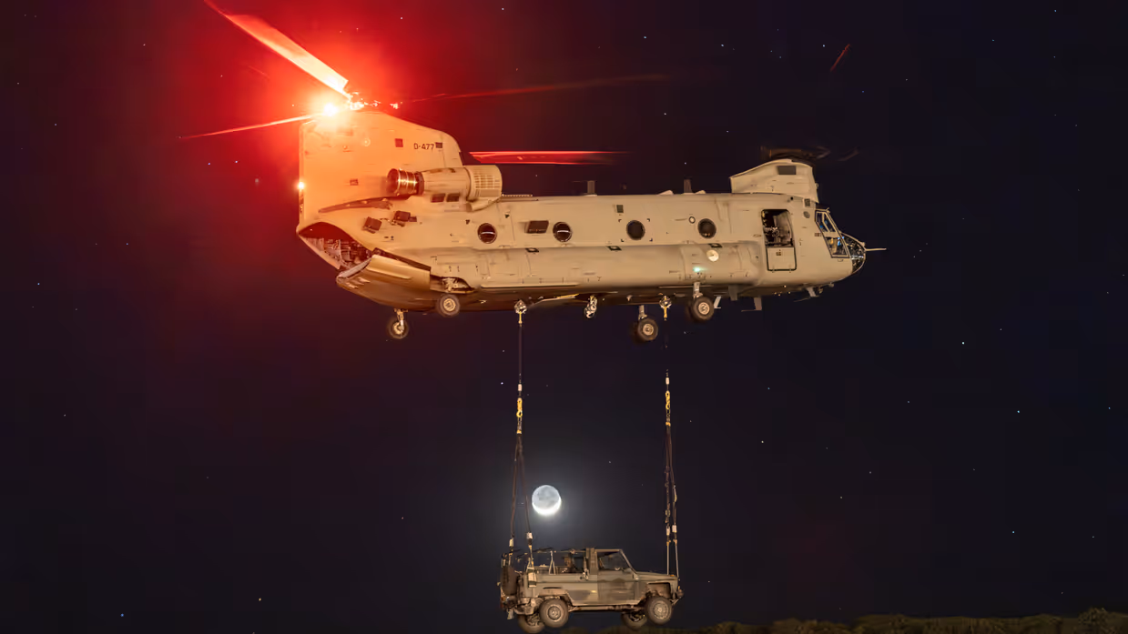 Een bij duisternis wegvliegende Chinook met eronder een militair voertuig aan kabels. Eronder begroeiing. Tussen de helikopter en het voertuig is de maan te zien.