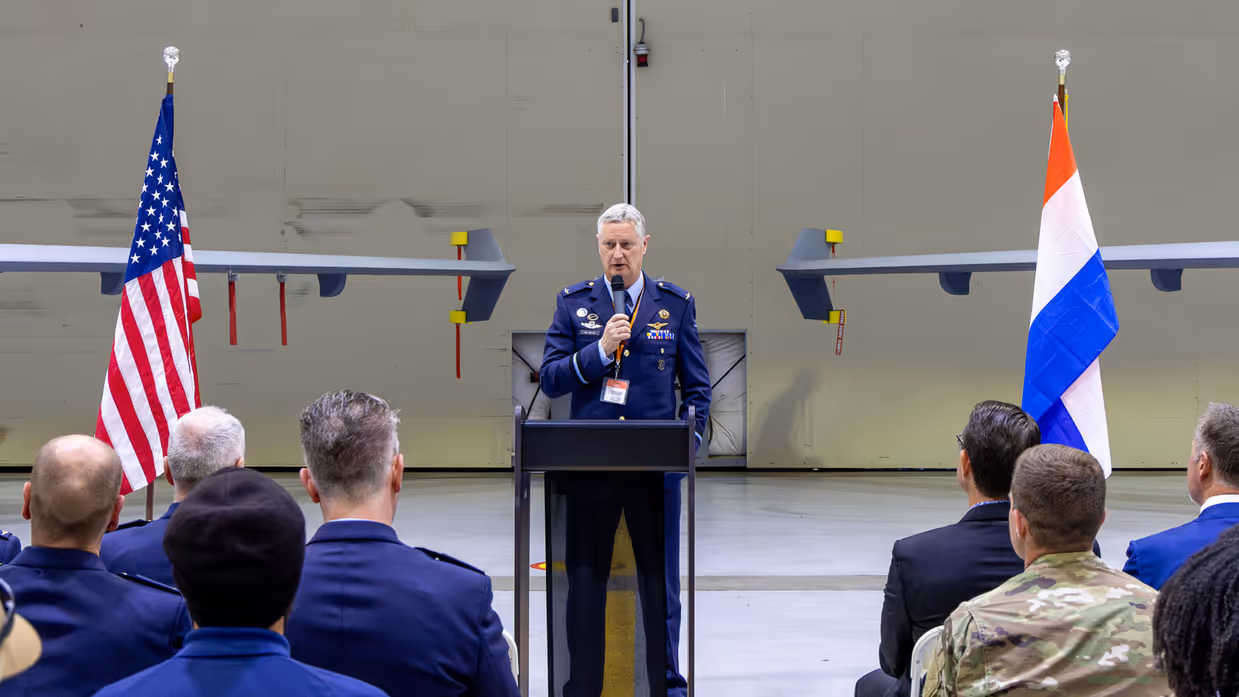Commodore Marcel van Egmond spreekt vanachter een spreekgestoelte publiek toe. Links en rechts achter hem een vleugel van twee MQ-9 Reapers. Voor de linkervleugel staat een Amerikaanse vlag, voor de rechter een Nederlandse.