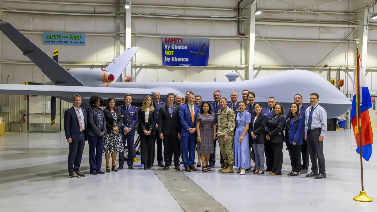 Een groep militairen en burgers poserend voor een MQ-9A Reaper in een hangaar.