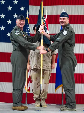 Voor een achtergrondvullende Amerikaanse vlag overhandigt Luijsterburg aan Van de Graaf een vlag. Beiden dragen een vliegeroveral en kijken lachend de camera in. Achter de vlag staat een militair in desert tenue.