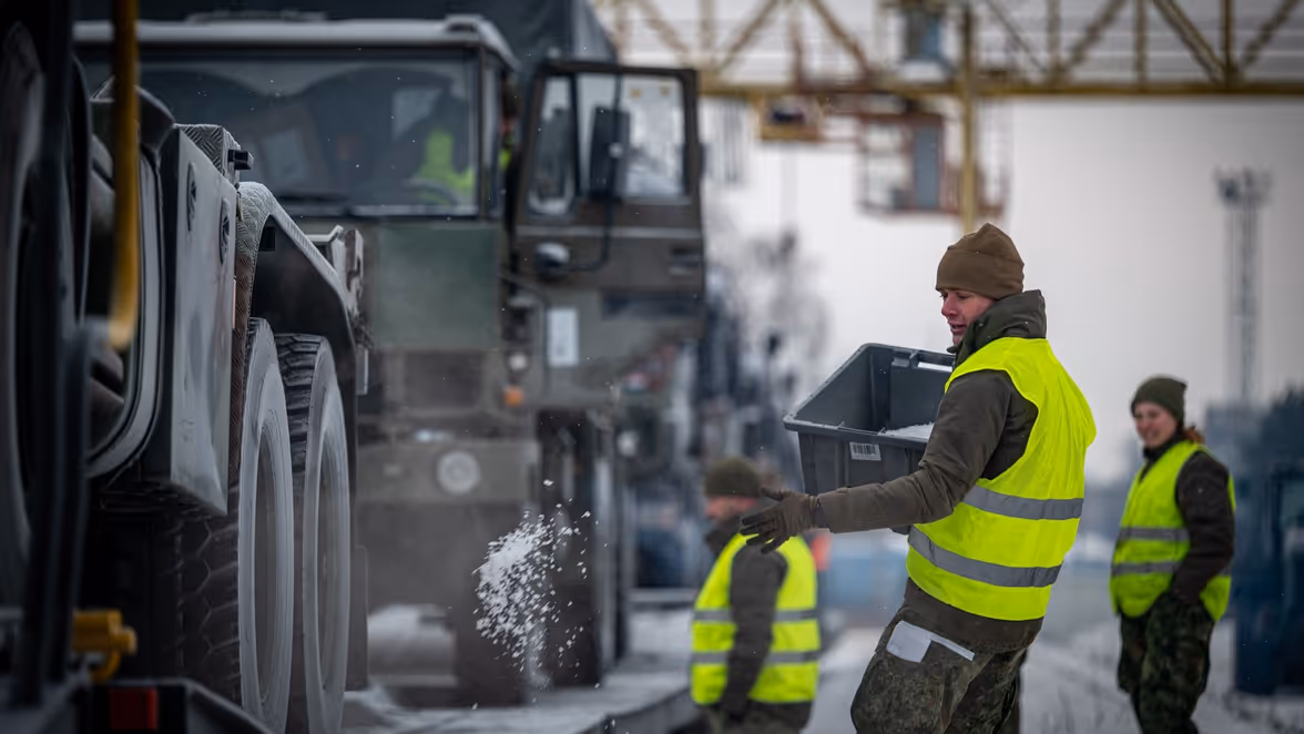 Drie personen in veiligheidshesjes en militaire kleding laden een grijze kist in een groot militair voertuig, terwijl sneeuw valt en de omgeving koud oogt.